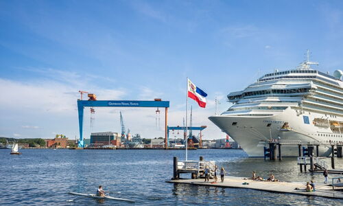 Kreuzfahrtschiff am Anleger in Kiel mit Blick auf die Werft German Naval Yards und Menschen am Steg bei sonnigem Wetter