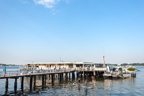 Steg mit Menschen und einem Gebäude am Ende, darüber blauer Himmel, darunter Wasser mit drei Paddlern