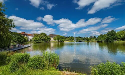 See mit Springbrunnen in einem Park, umgeben von Bäumen und Gebäuden unter blauem Himmel mit Wolken
